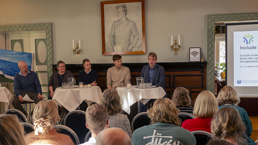 Forsamling i et rom lytter til et panel på fem personer, med et protrettmaleri av Fridtjof Nansen hengende over dem.