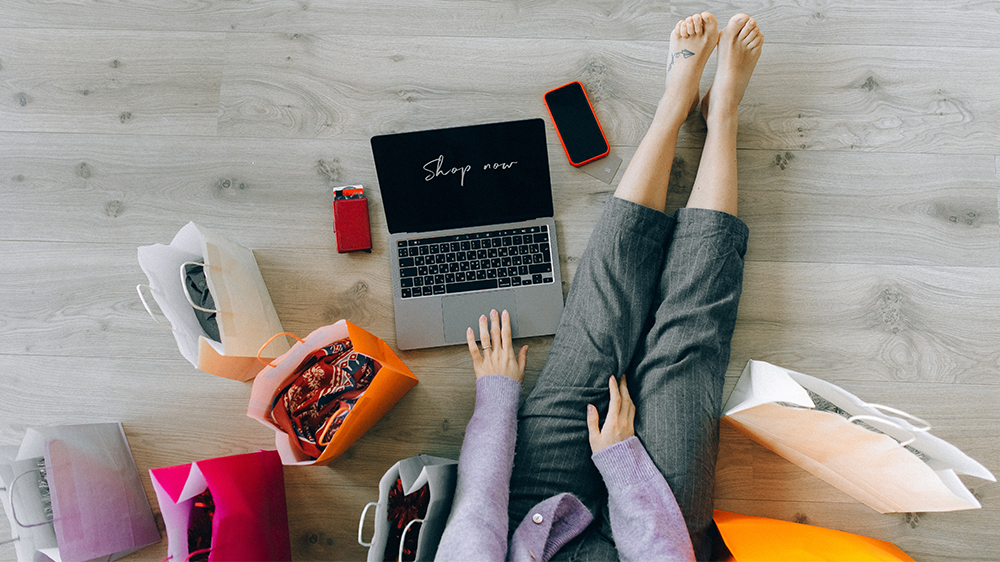 Sett ovenfra: person sitter på gulvet med handleposer og laptop der det står "shop now" på skjermen.